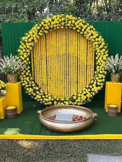A classic Haldi backdrop for outdoor functions. This setup features a large circular frame filled with strings of yellow marigolds, a green panel background, and a traditional brass urli in the center for the ceremony.