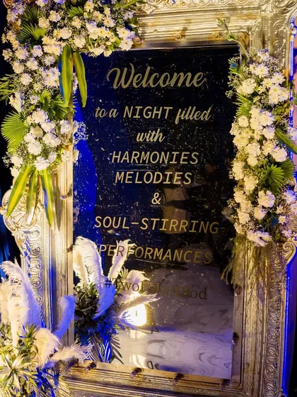 An elegant welcome sign for the Sangeet, written on a silver-framed mirror and adorned with white flowers, inviting guests to a "night filled with harmonies, melodies & soul-stirring performances."
