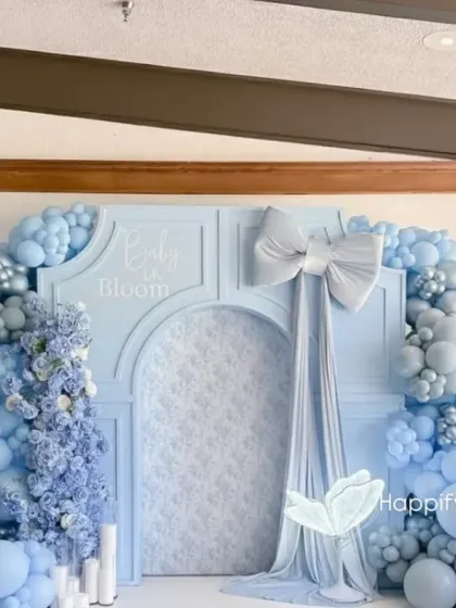 This premium "Baby in Bloom" baby shower backdrop features a sophisticated pastel blue theme. The decor includes a custom arched panel, a large fabric bow, and dense organic balloon garlands in varying shades of blue, accented with glowing butterfly props.