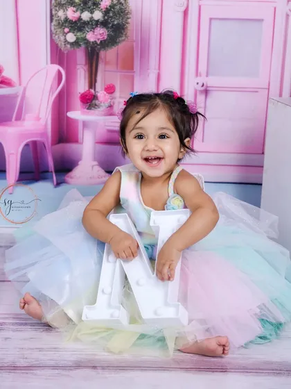 Holding the letter 'M', she shows off her playful side in a beautiful, multi-colored dress against a pink dollhouse backdrop.