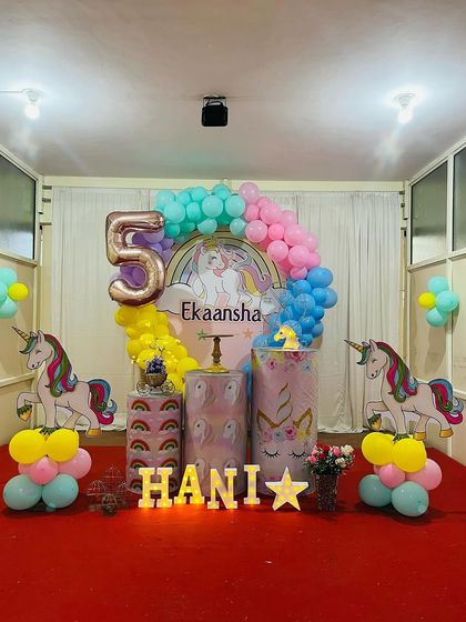 A classic unicorn theme for a 5th birthday. This setup includes a rainbow backdrop, unicorn cutouts, themed plinths, and a large foil '5' balloon.