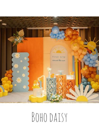 A 'Boho Daisy' themed party, also known as 'First Trip Around the Sun'. This wide shot shows the cheerful setup with orange and yellow tones, daisy props, and a rainbow.