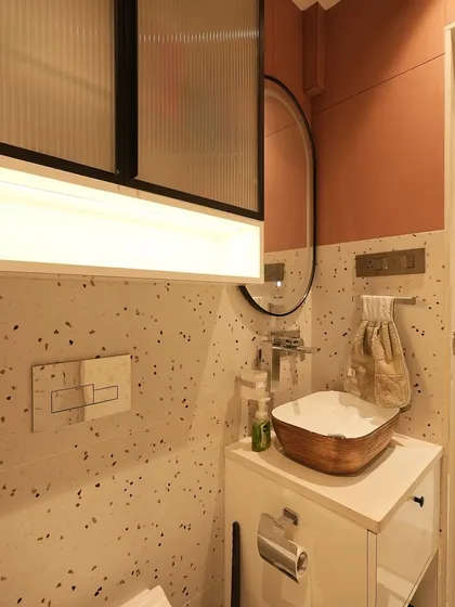 A compact and stylish powder room with terrazzo tiles, an oval mirror, and a unique wooden basin. The fluted glass detail on the overhead cabinet adds a touch of vintage charm.