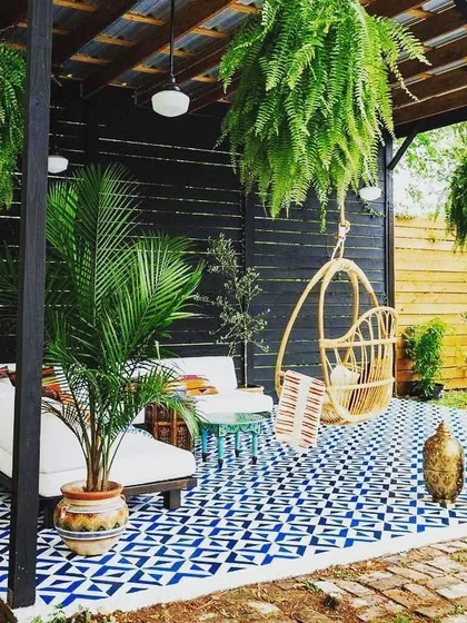 This covered patio design is all about boho-chic comfort. The space is defined by a bold patterned floor, a hanging swing chair, a plush daybed, and lots of hanging ferns, creating a relaxed and stylish outdoor living room.