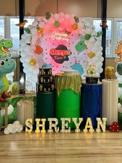 A grand dinosaur-themed setup for Shreyan's 6th birthday, with multiple dino cutouts and a white balloon arch with green accents.