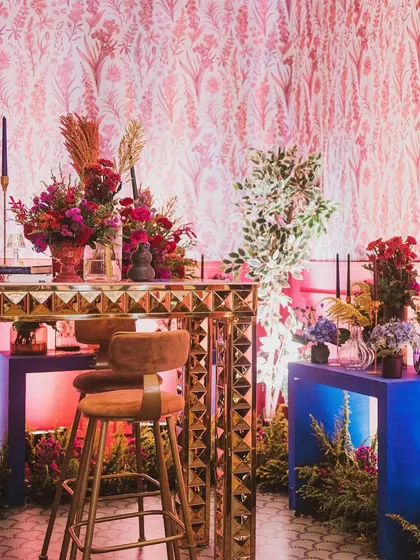 A stylish bar and lounge area designed for a modern sangeet. I paired a gold textured bar with bold blue consoles and vibrant red floral arrangements against a patterned pink wallpaper.
