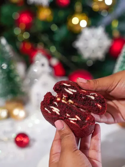 A gooey red velvet cookie with white chocolate chips, broken in half to show the soft center. The festive, out-of-focus background adds to the Christmas mood.