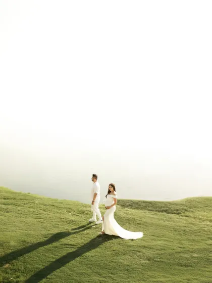 A stunning wide-angle shot of the couple on a grassy hill overlooking the water. The vast negative space and golden-hour shadows create a dramatic and minimalist composition.