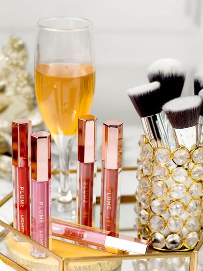 A collection of matte liquid lipsticks and makeup brushes are arranged in a crystal-studded holder, next to a glass of champagne. This setup creates a celebratory, glamorous vibe perfect for a beauty brand.
