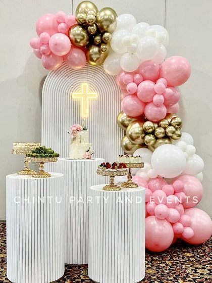 A beautiful baptism decoration for a baby girl, featuring a pink, white, and gold balloon arch. The setup includes a custom arched backdrop with a neon cross sign and fluted pedestals for desserts.