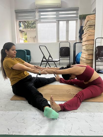An instructor assists a student in a deep seated forward bend, using her own body as leverage to provide a safe and effective stretch.