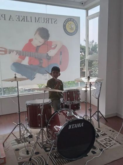 A young drummer sitting at his kit, ready to play. The poster in the back says "Strum Like a Pro".