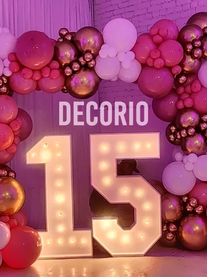 A vibrant 15th birthday party decoration with large, bright marquee numbers. The '15' is surrounded by a full arch of pink, white, and gold balloons, creating an exciting and photo-ready scene.