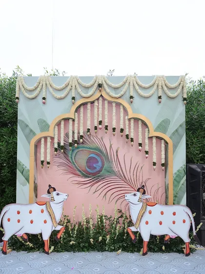 A unique and culturally rich photo booth backdrop for a Haldi ceremony. This setup features Pichwai-inspired art with cow motifs and a peacock feather, framed by traditional flower garlands.