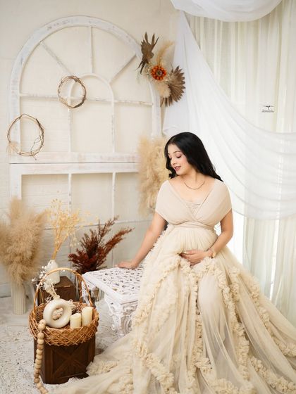 A full-length portrait of the mother-to-be, looking absolutely radiant in a flowing cream gown. The light and airy boho setup creates a dreamy, ethereal feel.