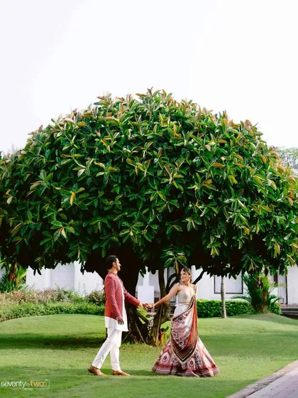 A wide shot of the couple holding hands under a magnificent, sprawling tree, showcasing the natural beauty of their Goan wedding venue.