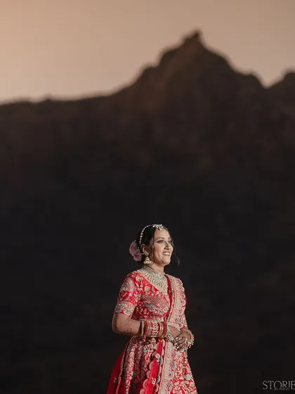 A bride in a stunning red lehenga stands against the majestic backdrop of a mountain in Rajasthan. Her joyful smile and the grand landscape come together to create a breathtaking destination wedding portrait.