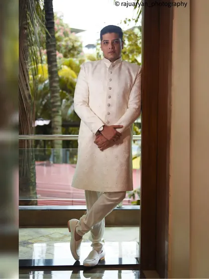 A sharp and stylish full-length portrait of a groom in a classic white sherwani. The natural light from the window and the confident pose create a sophisticated and timeless look for his wedding day.