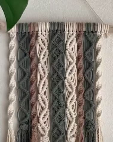 This wall hanging showcases a mix of colors and knotting styles, including the spiral "DNA" knot. The combination of off-white, grey, and dusty rose creates a soft, modern look.