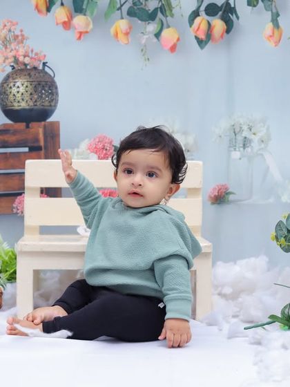 A sweet toddler in a green hoodie sits on a small white bench amidst a sea of clouds. The floral background adds a touch of spring to this dreamy scene.