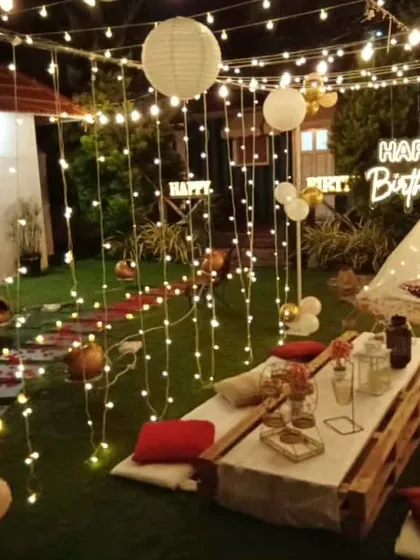 A full view of an elaborate backyard birthday party. The space is filled with string lights and paper lanterns, illuminating a long boho picnic table and a cozy teepee tent in the background.