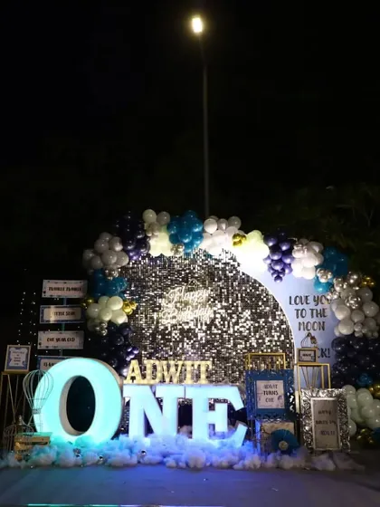 A "Love You to the Moon & Back" themed first birthday setup. The decor features a silver sequin backdrop, a large light-up "ONE" sign, and a blue and silver balloon arch.