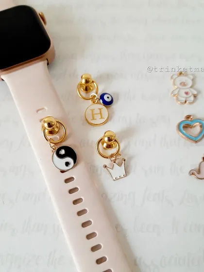 A closer look at the smartwatch charms, including a yin-yang, a crown, and a personalized initial 'H' with an evil eye, attached to a pink watch band.