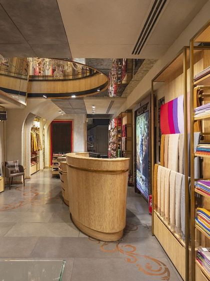 The main retail floor at Ahujasons features a curved wooden counter and brass-framed shelving. The design uses soft, organic forms and a neutral palette to create a calm and sophisticated canvas for the vibrant, handcrafted textiles on display.