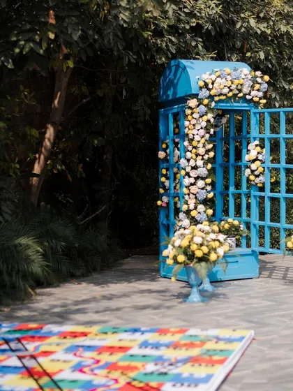 A quirky photo-op featuring a vintage British telephone booth, painted bright blue and overflowing with flowers, set next to a giant snakes and ladders game board.