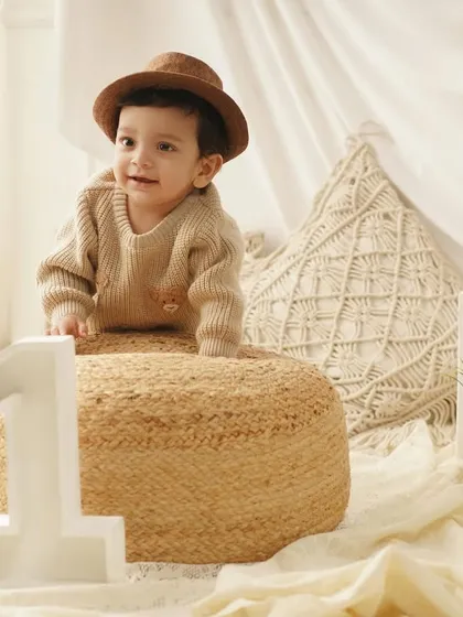 A dapper little boy in a hat and sweater poses with a '1' prop for his first birthday shoot, set against a warm, neutral-toned boho backdrop.