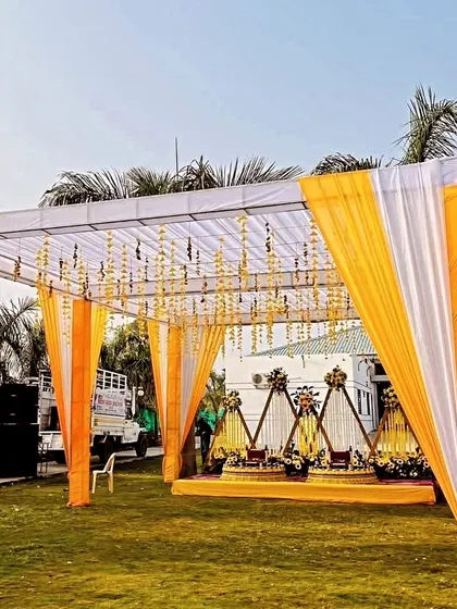 A wide view of an outdoor Haldi event, with a large canopy decorated in white and yellow drapes, perfect for a sunny day.