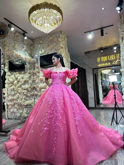A full view of our gorgeous glittery rose pink engagement ball gown. The dramatic off-shoulder ruffles and vine-like embroidery create a look that is both bold and romantic. The studio's flower wall provides the perfect backdrop.