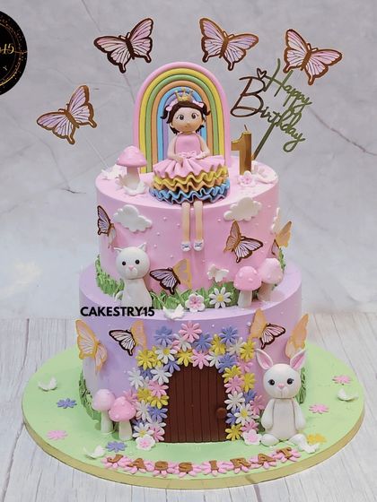 A dreamy two-tier rainbow doll cake for a first birthday. This cake, in black forest and pineapple flavors, features a doll topper, a rainbow, and a whimsical garden with a bunny and a kitten.