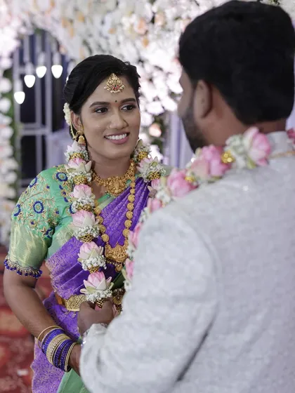 A sweet moment between bride Divya and groom Pavan at their engagement. Divya's look is fresh and vibrant, with a purple and green saree and makeup that highlights her happy glow.
