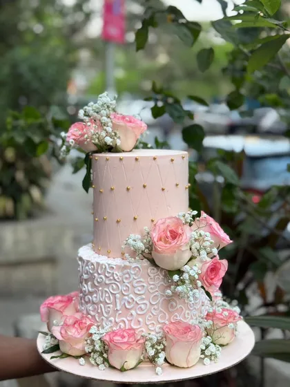 An elegant two-tier engagement cake in soft pink. The bottom tier features delicate white piping, while the top has a simple dotted pattern, all brought together with beautiful fresh pink roses.