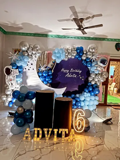 An out-of-this-world space-themed birthday decoration. This setup features a deep blue and silver balloon arch, a crescent moon cutout, and astronaut props to create a galactic adventure for the birthday star.