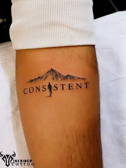 A motivational tattoo that integrates a mountain range and a lone hiker into the word "CONSISTENT." It’s a powerful reminder that every step forward counts.