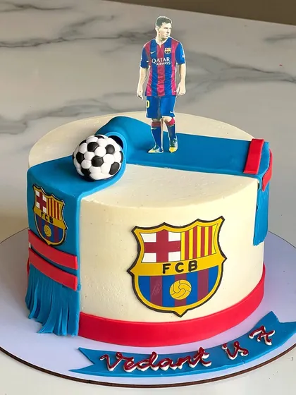 For the little football fan, this cake is themed around FC Barcelona. It features the team's logo and a scarf draped over the cake, complete with a fondant soccer ball and a player cutout.