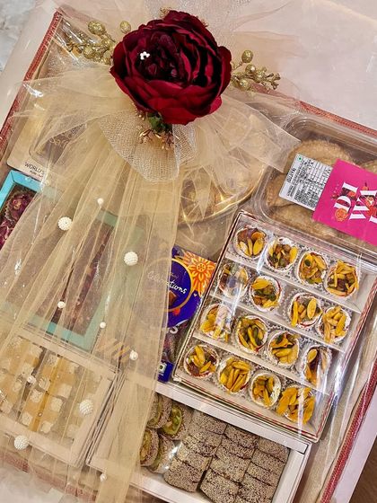 A close-up of a festive gift box filled with a variety of Indian sweets (mithai). The box is wrapped in sheer net with a beautiful red rose on top, making it an elegant gift for any wedding ceremony.
