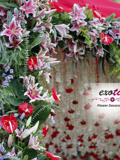 A close-up of a floral wall with red lilies and anthuriums. The background features a curtain of hanging red flower buds, adding depth and texture to the design.