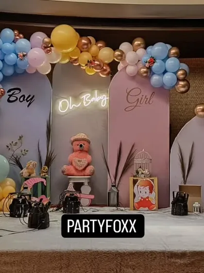 A "Boy or Girl" gender reveal party setup with pink and blue arched backdrops, a central "Oh Baby" neon sign, and a teddy bear prop.
