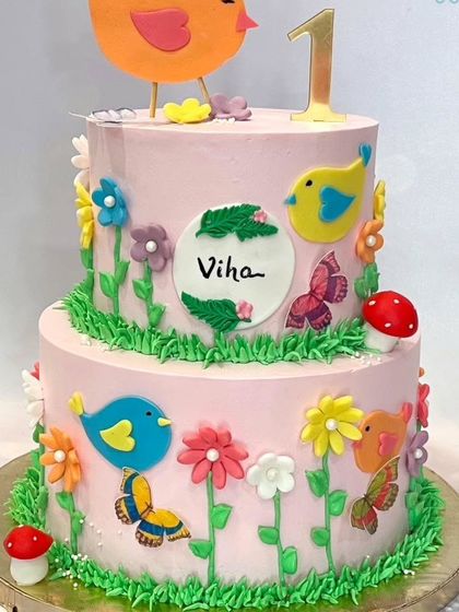 A closer view of the birds and garden themed two-tier cake, showing the detailed fondant work and vibrant colors on a pink whipped cream base.