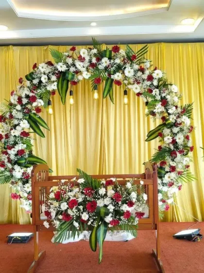 A close-up of the cradle decoration for a naming ceremony. The wooden cradle is adorned with red and white flowers, matching the large floral arch in the background.