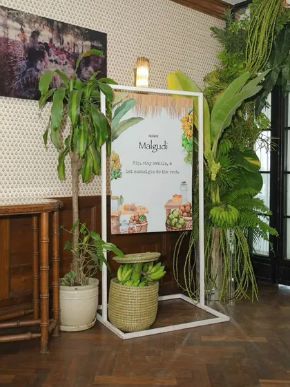 The welcome sign for the 'Malgudi' collection launch, framed by a lush arch of tropical greenery.