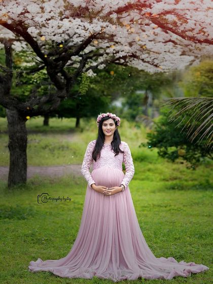 A serene solo portrait of a mother-to-be in a pink lace gown and floral crown, standing under a tree with white blossoms.