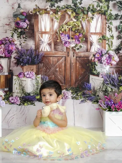Holding a tiny teddy bear, this little one looks perfectly at home in her magical garden, surrounded by lush greenery and purple blooms.
