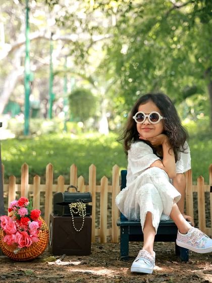 A stylish little girl posing confidently in a park. The natural setting with a rustic fence and baskets of flowers creates a charming scene for this outdoor fashion shoot.