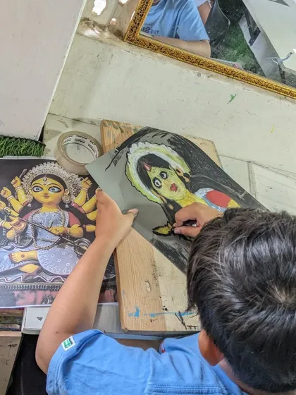 An overhead view of a student working on a detailed drawing of Durga Ma, using a reference image to capture the traditional features.