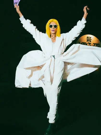 A moment of pure expression from a high-fashion night shoot. The model's arms are raised in a gesture of freedom, the flowing white coat creating a dramatic silhouette against the dark background.