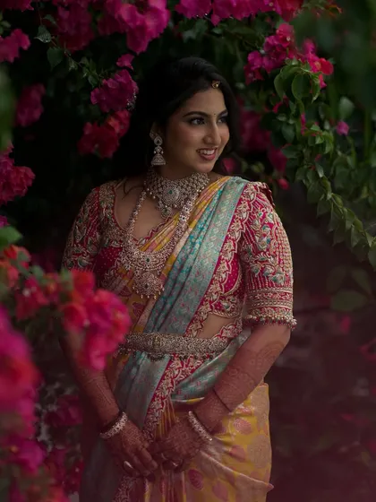 A radiant bride captured amidst a backdrop of vibrant bougainvillea. Her custom-designed saree features a unique color palette, paired with a heavily embellished blouse that combines traditional patterns with a modern aesthetic for her special day.